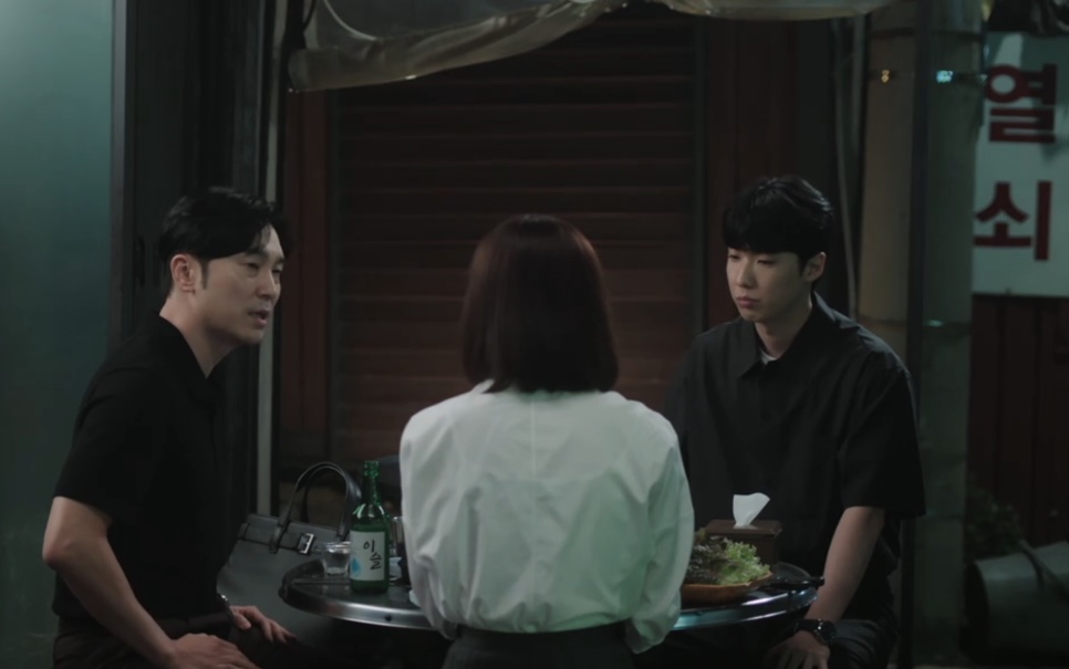 Park Ki-tae sentado a uma mesa na rua, à noite, com dois colegas de trabalho, em The Ordinary Jackpot
