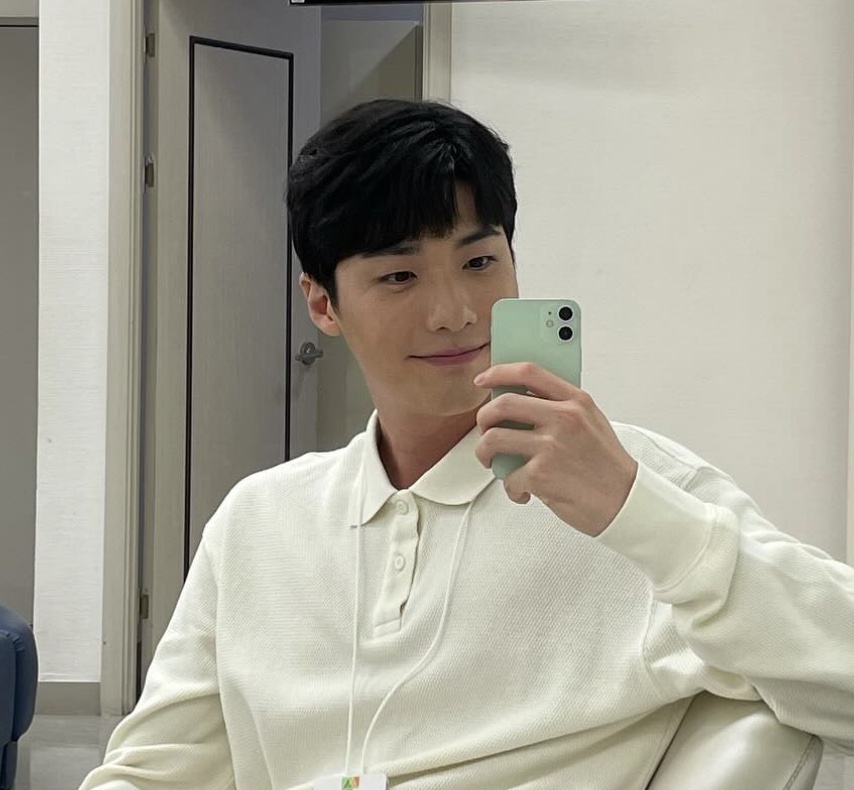 O ator Hong Jin-gi de camisa polo clara e crachá, tirando selfie no espelho