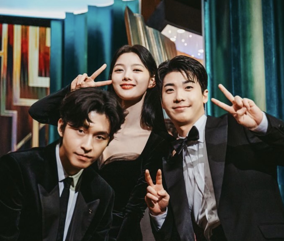 O ator Hong Jin-gi de terno e gravata-borboleta, fazendo sinal de V com dois colegas de elenco