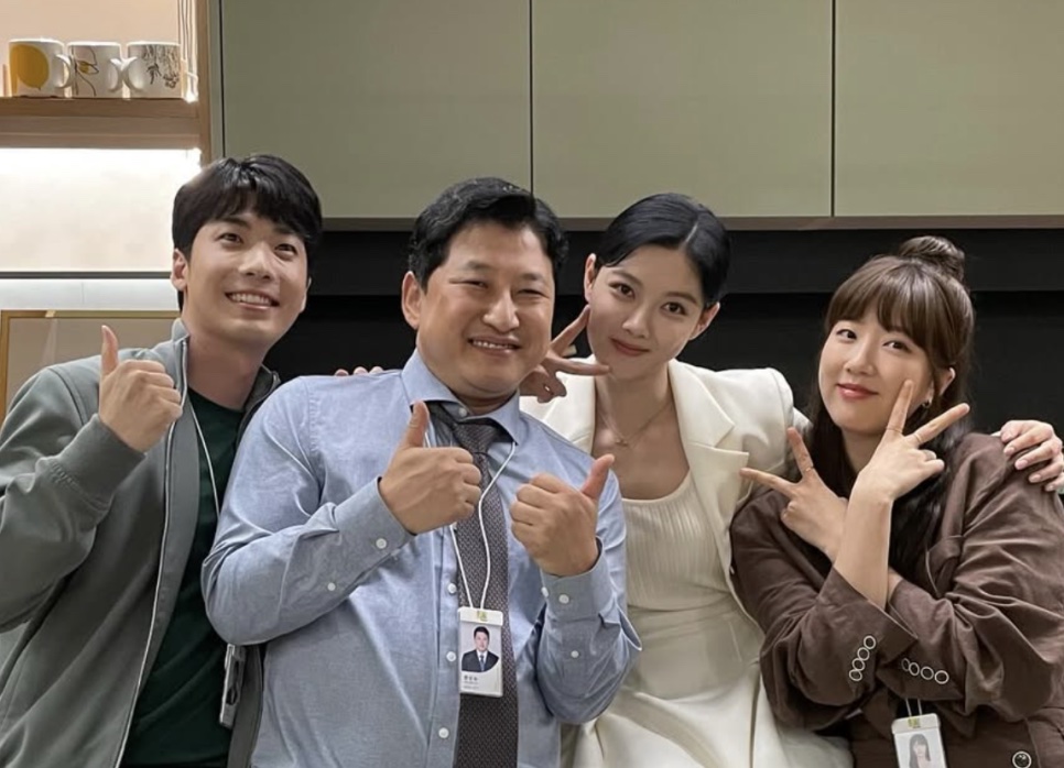 O ator Hong Jin-gi fazendo sinal de positivo com colegas de elenco num cenário de escritório