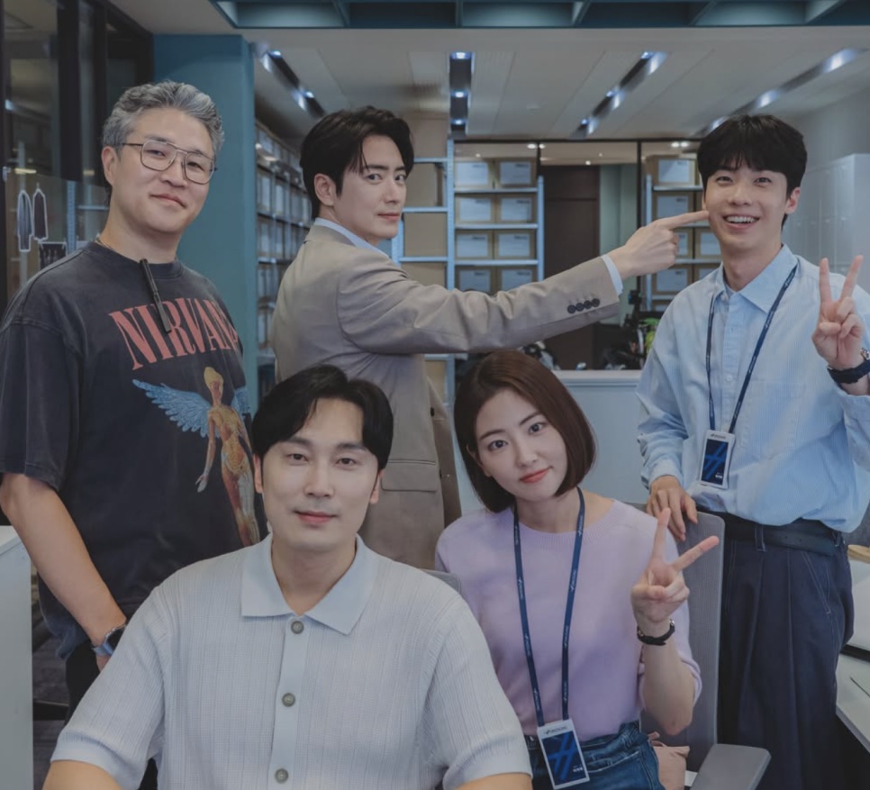 O ator Hong Jin-gi fazendo sinal de V com colegas de elenco de The Ordinary Jackpot num cenário de escritório