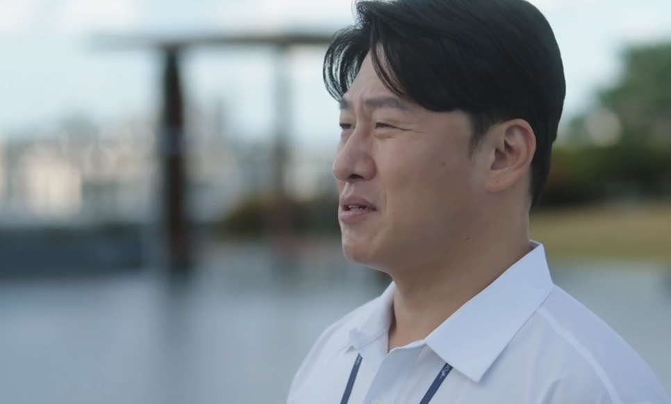 Oh Dae-hwan de camisa branca e crachá, sorrindo num terraço, no papel do Jung Seon-hyuk em The Ordinary Jackpot