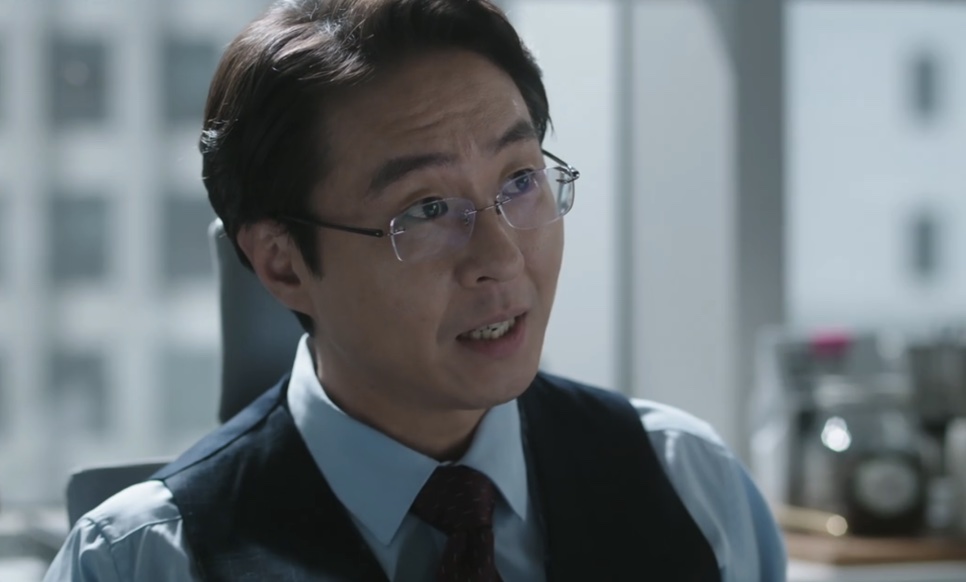 Lee Hyun-kyun de óculos e colete escuro, falando no escritório, no papel do Jang Kyung-chul em The Ordinary Jackpot