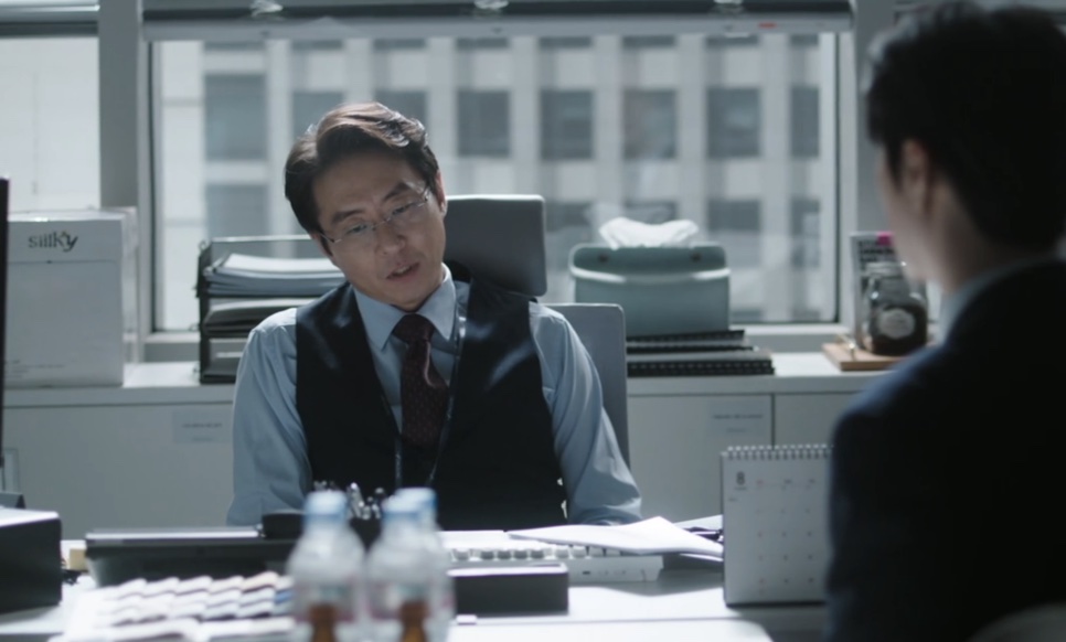 Jang Kyung-chul sentado à mesa dele no escritório, conversando com alguém de costas, em The Ordinary Jackpot