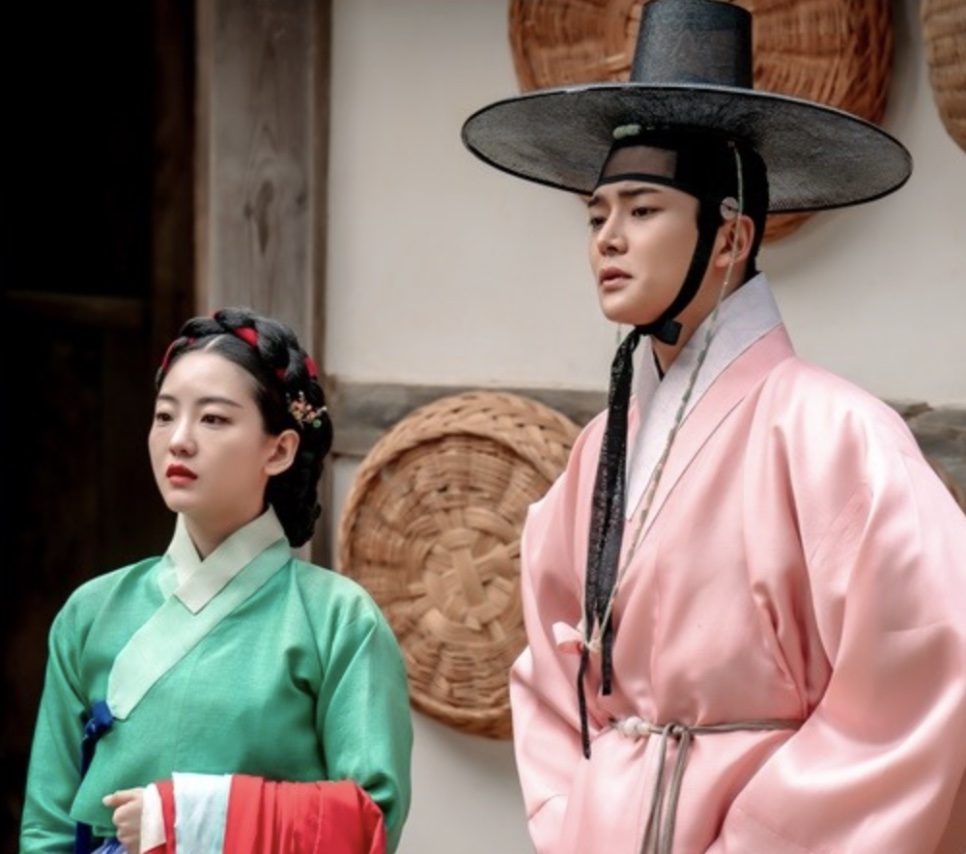 Um rapaz de hanbok rosa e chapéu tradicional coreano ao lado de uma moça de hanbok verde, numa cena de The Matchmakers