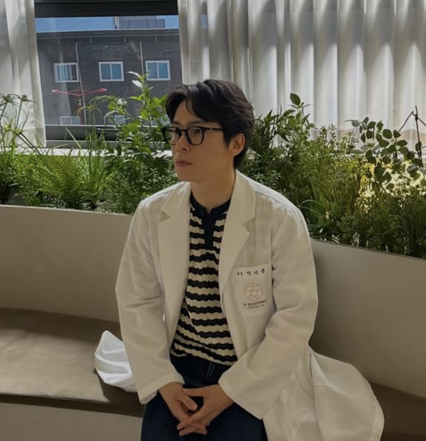 Lee Jae-won de jaleco branco e óculos, sentado ao ar livre