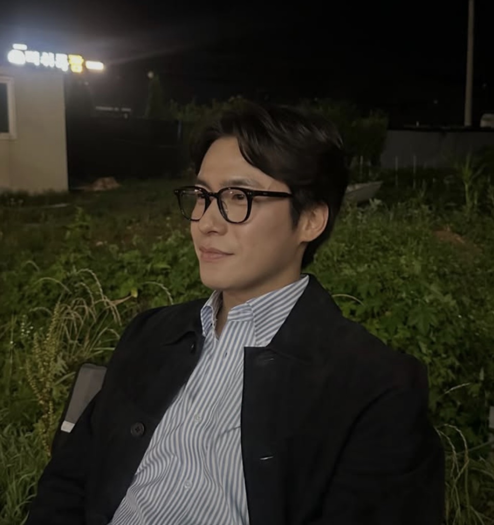 Lee Jae-won de óculos e casaco escuro, olhando de lado, em foto noturna