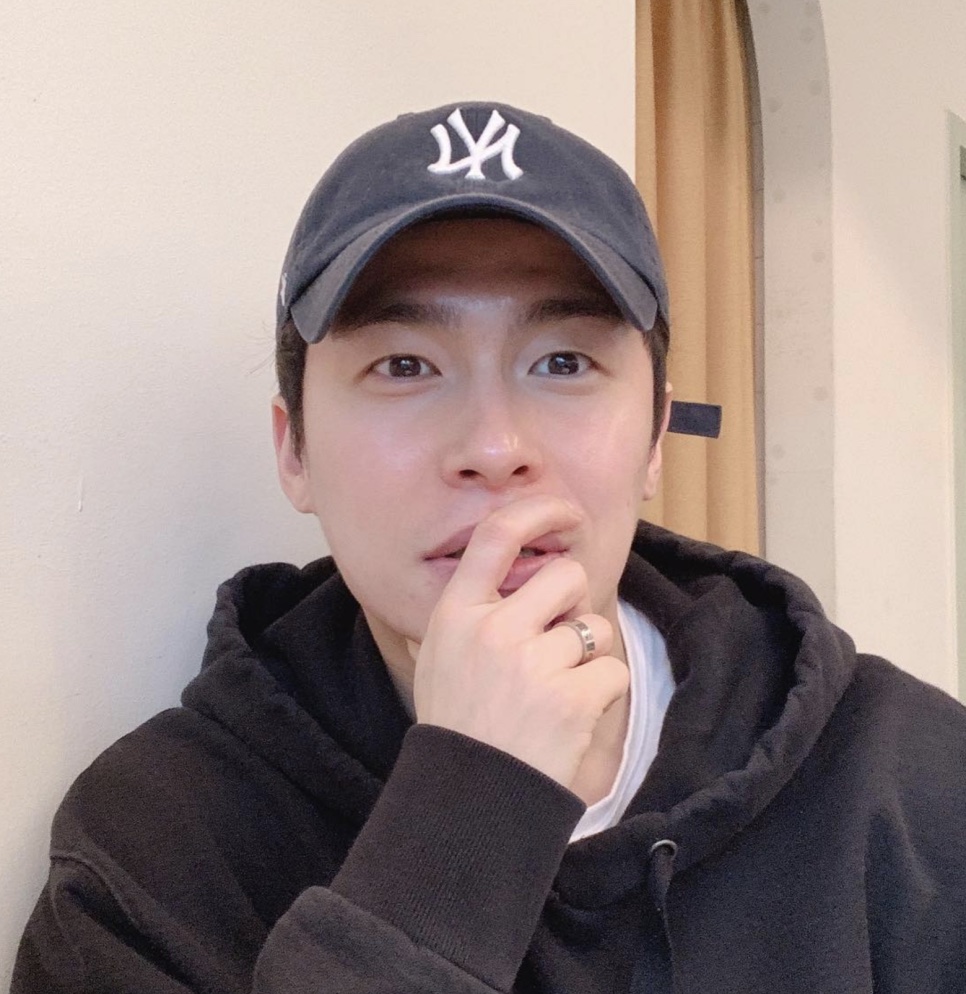 Lee Jae-won de boné e moletom, com a mão perto do rosto, em selfie