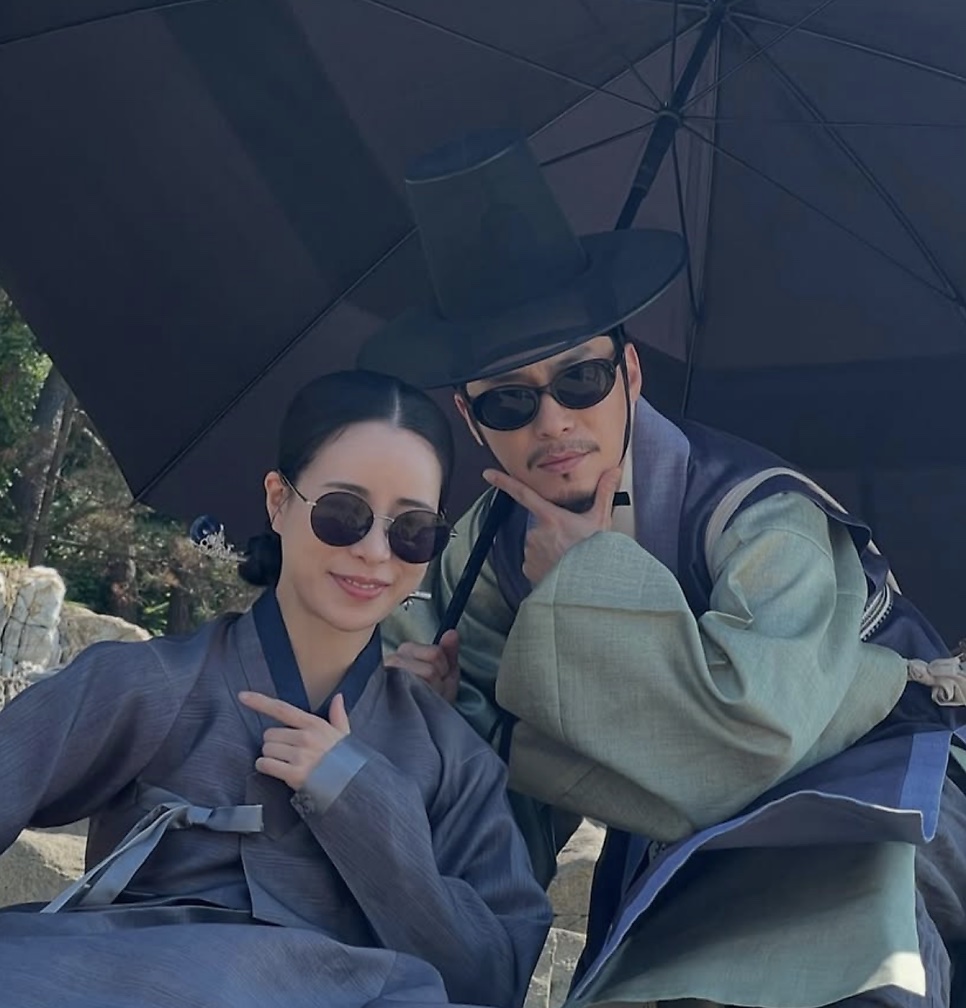 Lee Jae-won de hanbok (traje tradicional coreano) e chapéu preto, ao lado de uma atriz, em gravação de um dorama de época