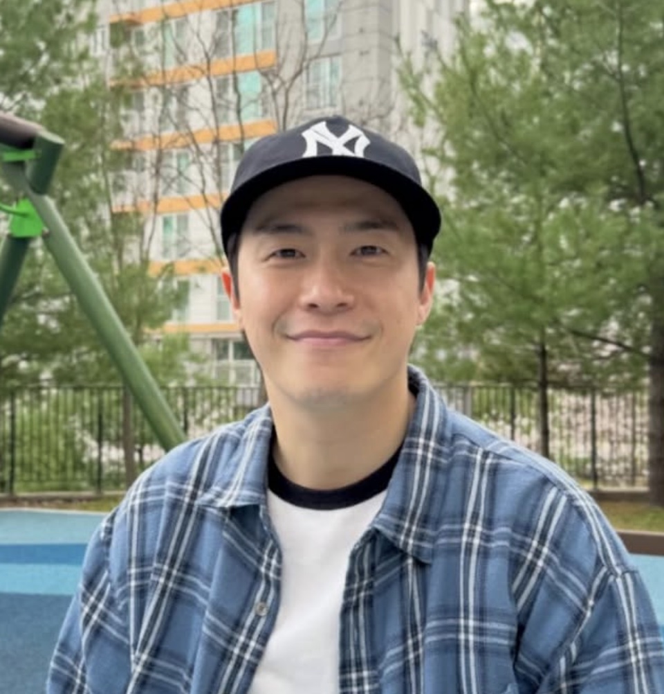 Lee Jae-won de boné e camisa xadrez, sorrindo em um parquinho