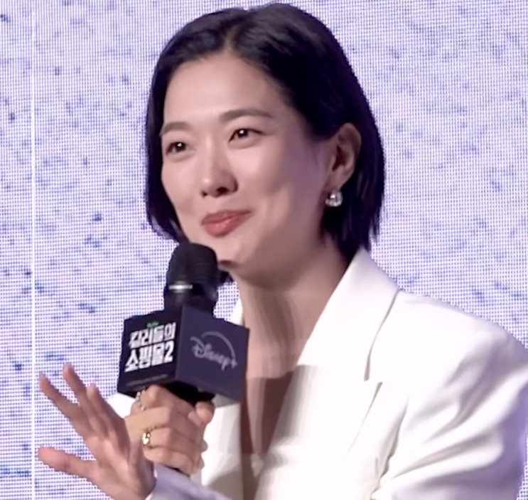 Jung Yun-ha de blazer branco falando ao microfone numa coletiva de imprensa