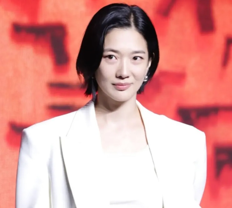 Jung Yun-ha de terno branco diante de um fundo vermelho, olhando para o lado