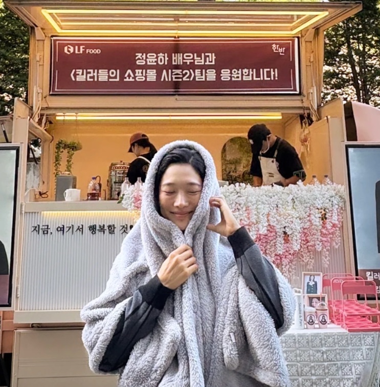 Jung Yun-ha enrolada num cobertor diante de um food truck enviado por fãs para a equipe da série
