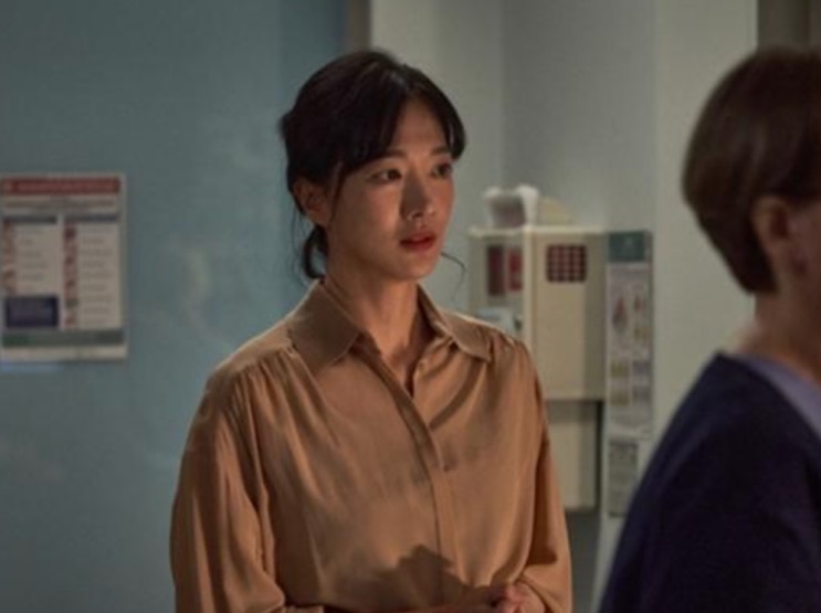 Jung Yun-ha em cena de Exhuma, de blusa marrom, conversando com outra pessoa