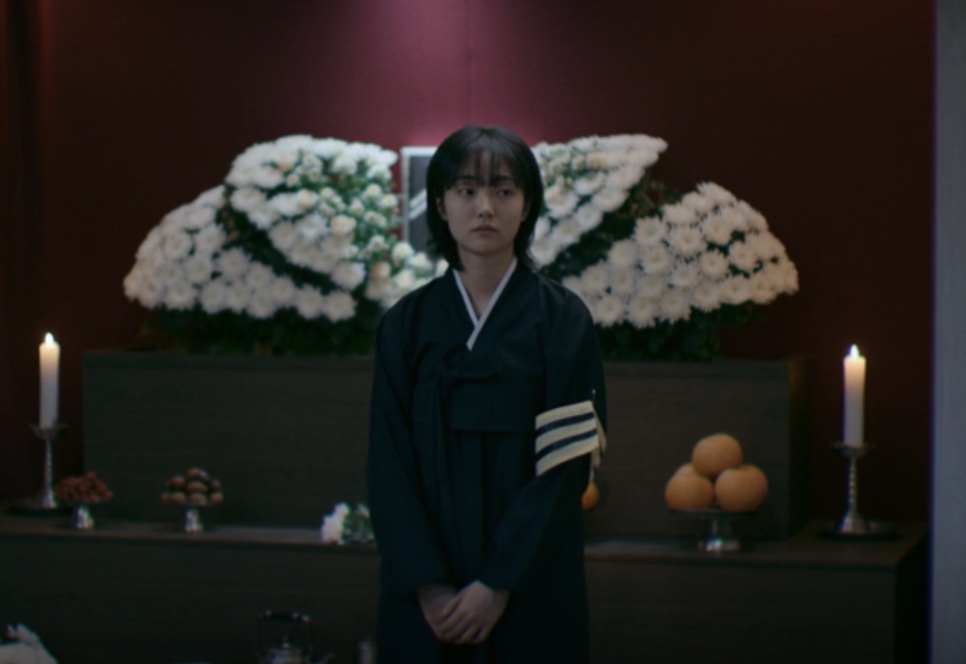 Jeong Ji-an de luto, sentada diante do altar com a foto do tio e coroas de flores brancas