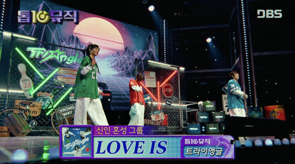 O Triangle cantando 'Love Is' no palco de um programa musical, com o nome da música no letreiro, em Wild Sing