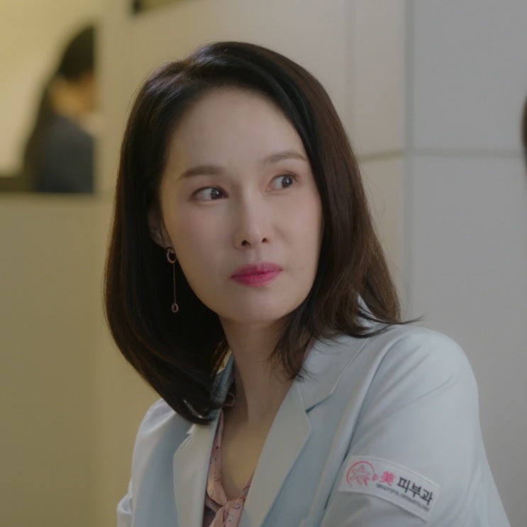 Baek Mi-hee de jaleco branco, sorrindo de leve em um corredor de hospital