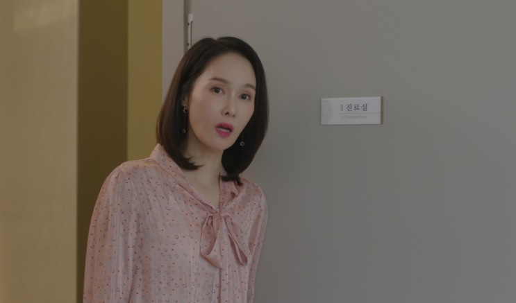 Baek Mi-hee de blusa rosa, de pé em um corredor claro