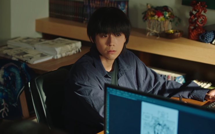 Shin Dae-ryuk de cabelo escuro, sentado diante de um computador em uma sala bagunçada