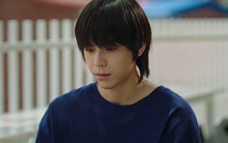 Shin Dae-ryuk de camiseta azul, olhando para baixo em um ambiente externo